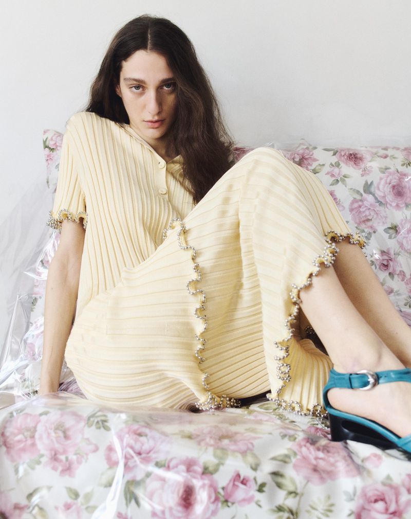 A young adult woman with long dark hair is sitting on a bed with a floral pattern, wearing a yellow ribbed knit dress and blue sandals.