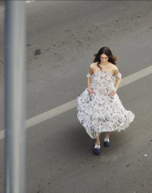 Overhead shot of a woman in an off-the-shoulder floral dress and navy shoes walking down a grey asphalt street.