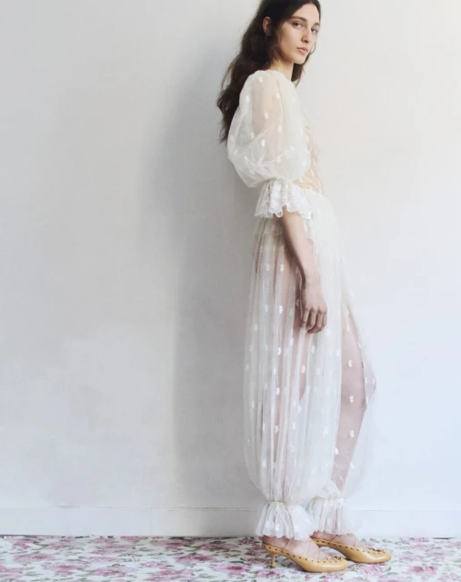 A woman in a light, translucent dress with polka dots poses against a white wall, with a floral rug adding a touch of color.