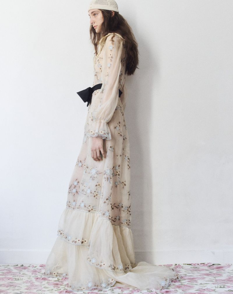 An adult woman wears a long-sleeved, floral-patterned dress, black bow, and a white crocheted hat against a white wall with floral flooring.