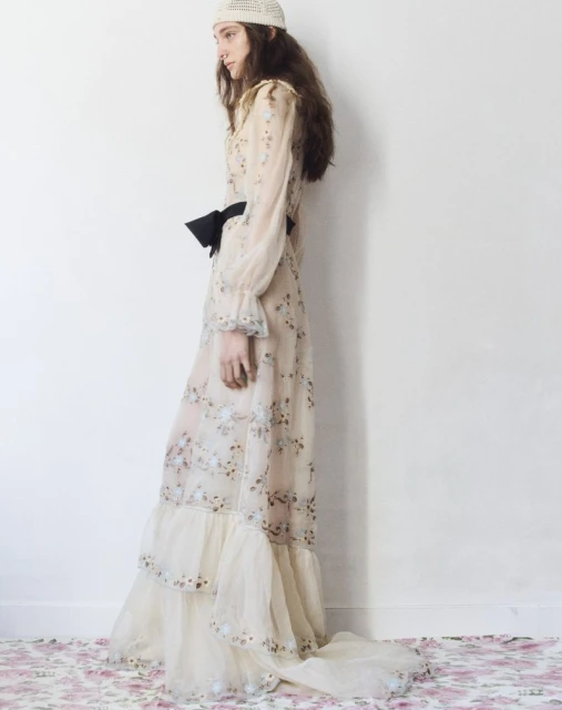An adult woman wears a long-sleeved, floral-patterned dress, black bow, and a white crocheted hat against a white wall with floral flooring.