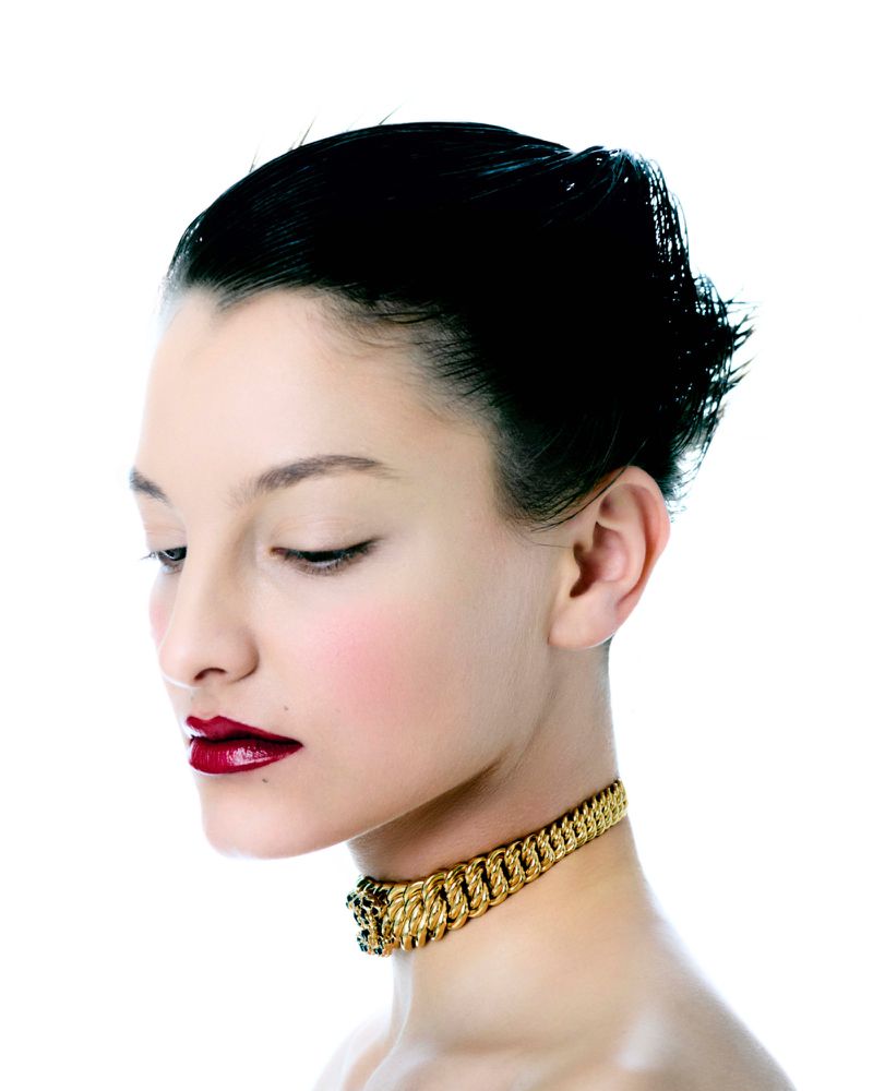 A headshot of an adult female with dark hair in an updo, wearing a thick gold choker and dark red lipstick, set against a stark white background.
