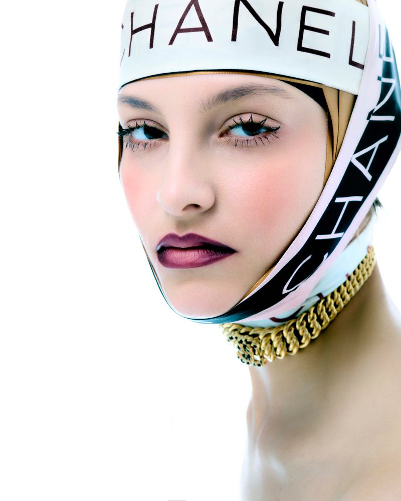 Close-up portrait of a young woman with dark lipstick and eye makeup, wearing a Chanel scarf and gold chain necklace, set against a bright white backdrop.