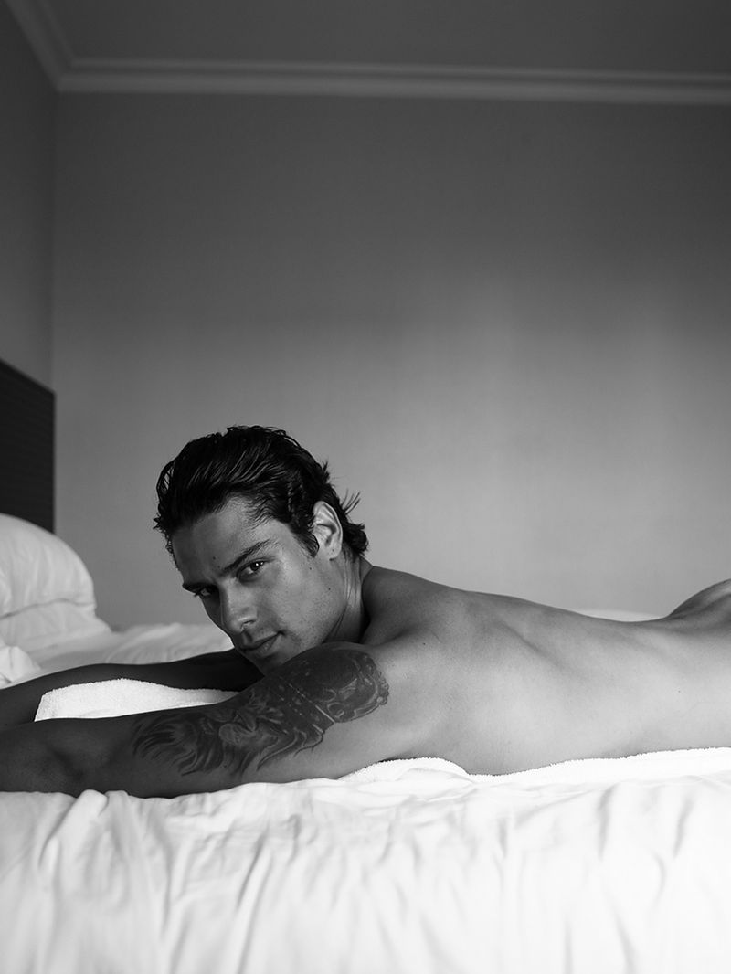 Black and white portrait of a shirtless adult male lying on a white bed with a tattoo on his arm, looking at the camera.