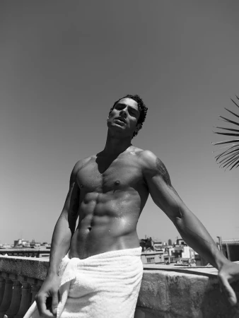 Black and white portrait of a muscular shirtless adult male wearing a towel, posing against a rooftop with a clear sky backdrop.