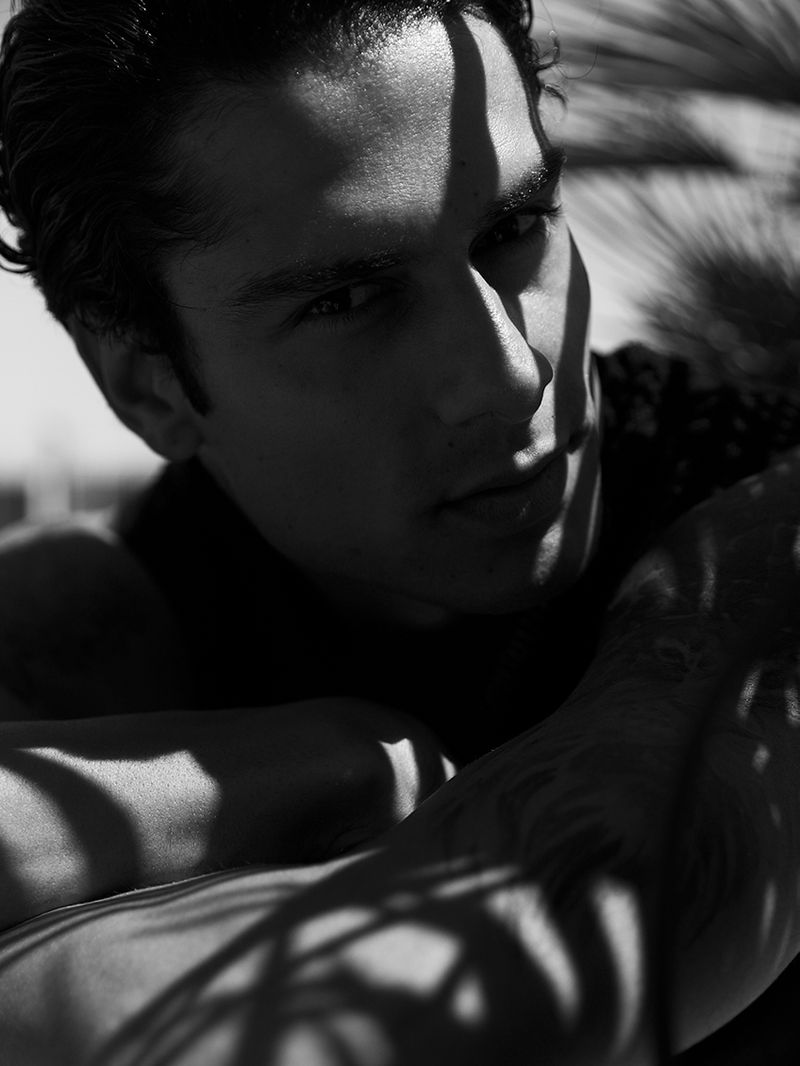 Black and white portrait of a man with shadows from palm fronds falling across his face and body, creating a high-contrast tropical mood.