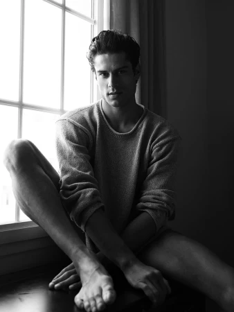 Black and white portrait of a young adult man sitting by a window in a sweater, looking at the camera with a neutral expression.