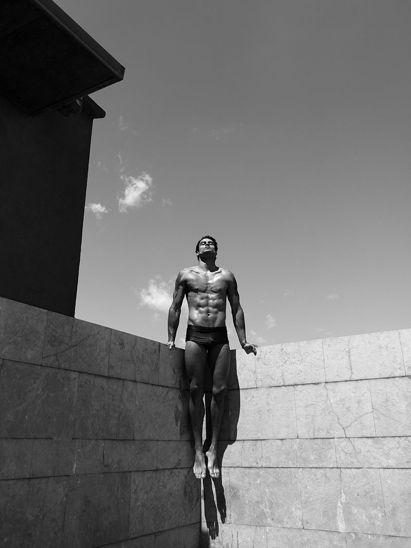 Monochrome shot of a muscular man posing on a building ledge against a bright sky, showcasing fitness and architecture.