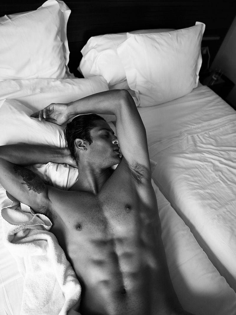 An adult man is lying on white bedsheets with his arms behind his head, creating a relaxed and intimate monochrome portrait.