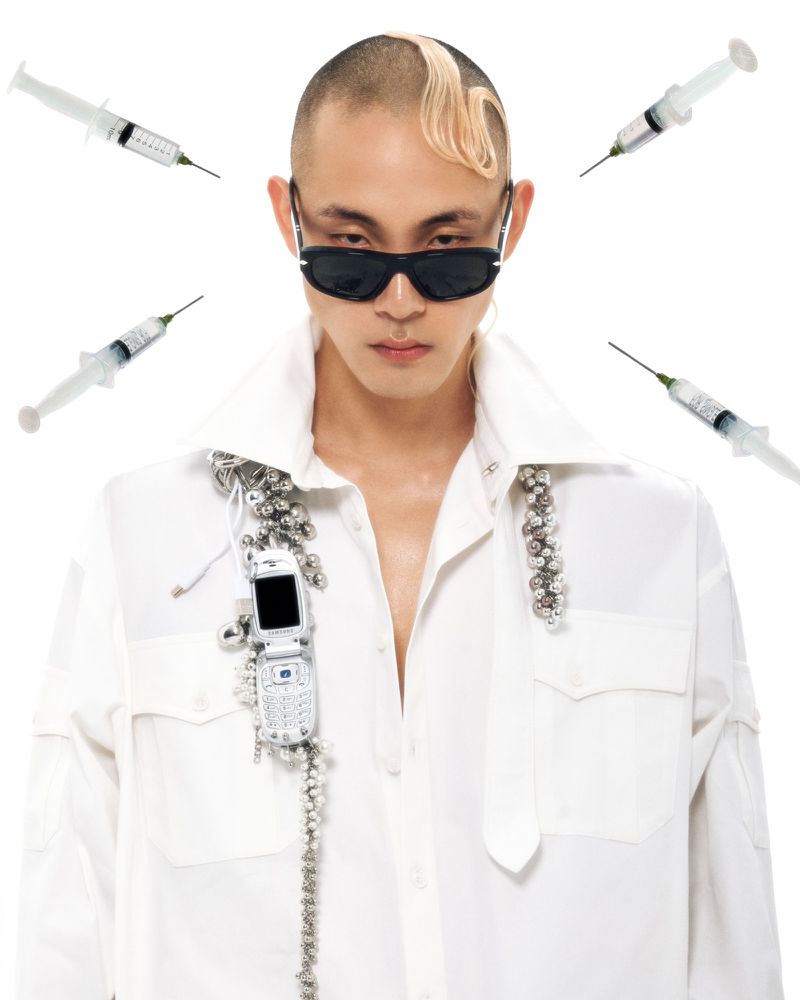 Portrait of a person with a shaved head wearing sunglasses and a white shirt, accessorized with repurposed flip phone on a white backdrop.