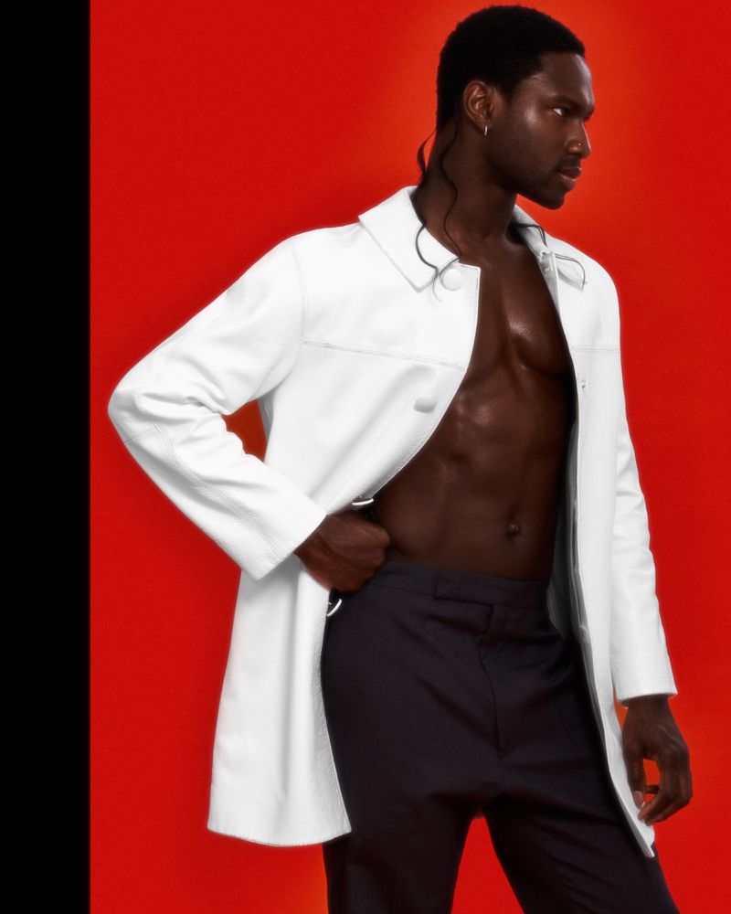 A dark-skinned male model wearing a white trench coat with no shirt stands in front of a solid red background in a studio setting.