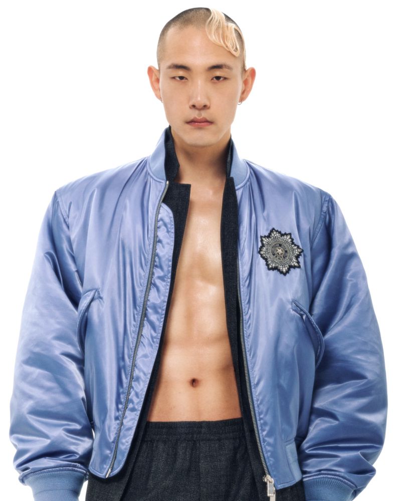 Man in light blue bomber jacket with abs visible in studio shot. Model poses against white backdrop in fashion style.