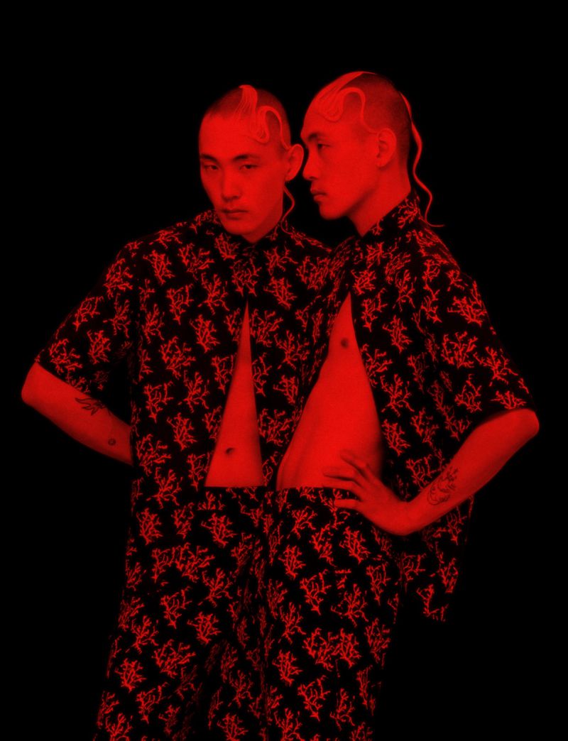 Two people in matching red and black patterned outfits stand closely against a solid black background with red lighting in studio.