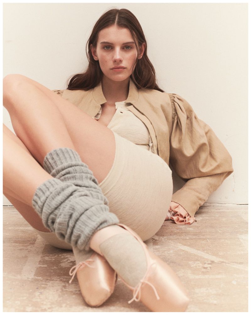 A young woman in a light jacket and bodysuit poses indoors wearing ballet shoes and leg warmers in a fashion editorial-style photo.