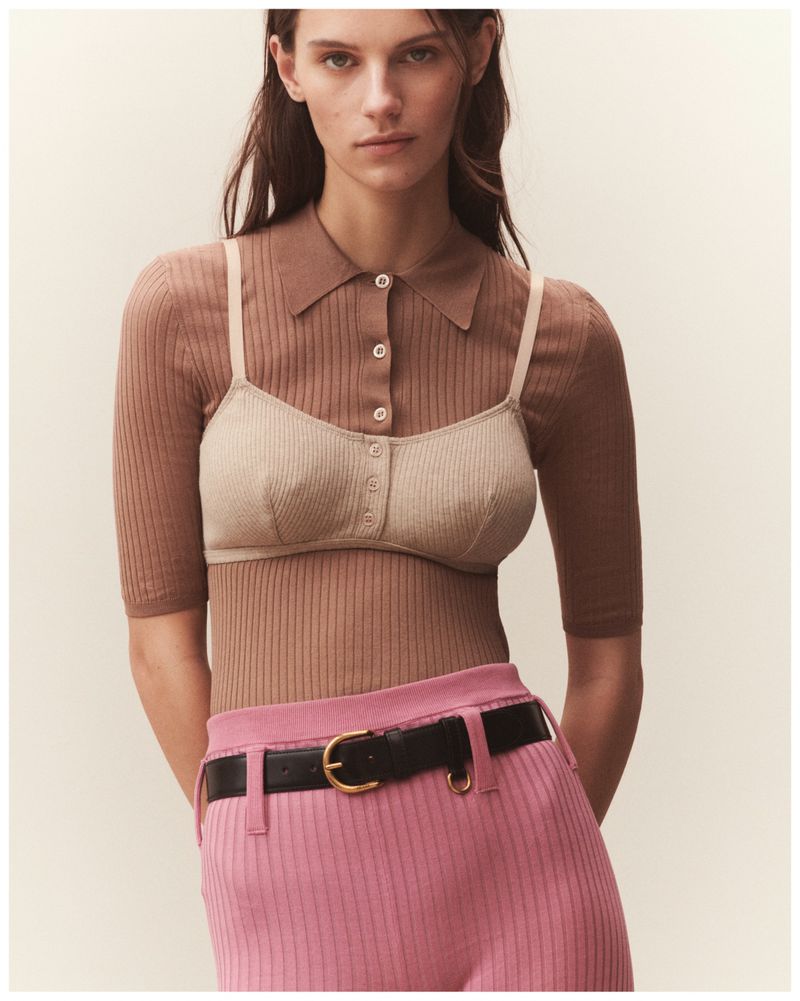 Brunette woman in beige bra top, tan collared shirt, and pink pants with black belt posing in a medium shot against a neutral background.