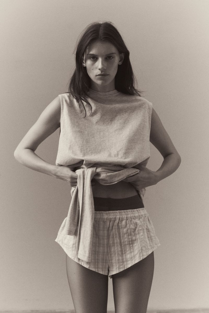 Woman in sleeveless top and striped shorts poses indoors with hands lifting her shirt, neutral color palette, and natural lighting.