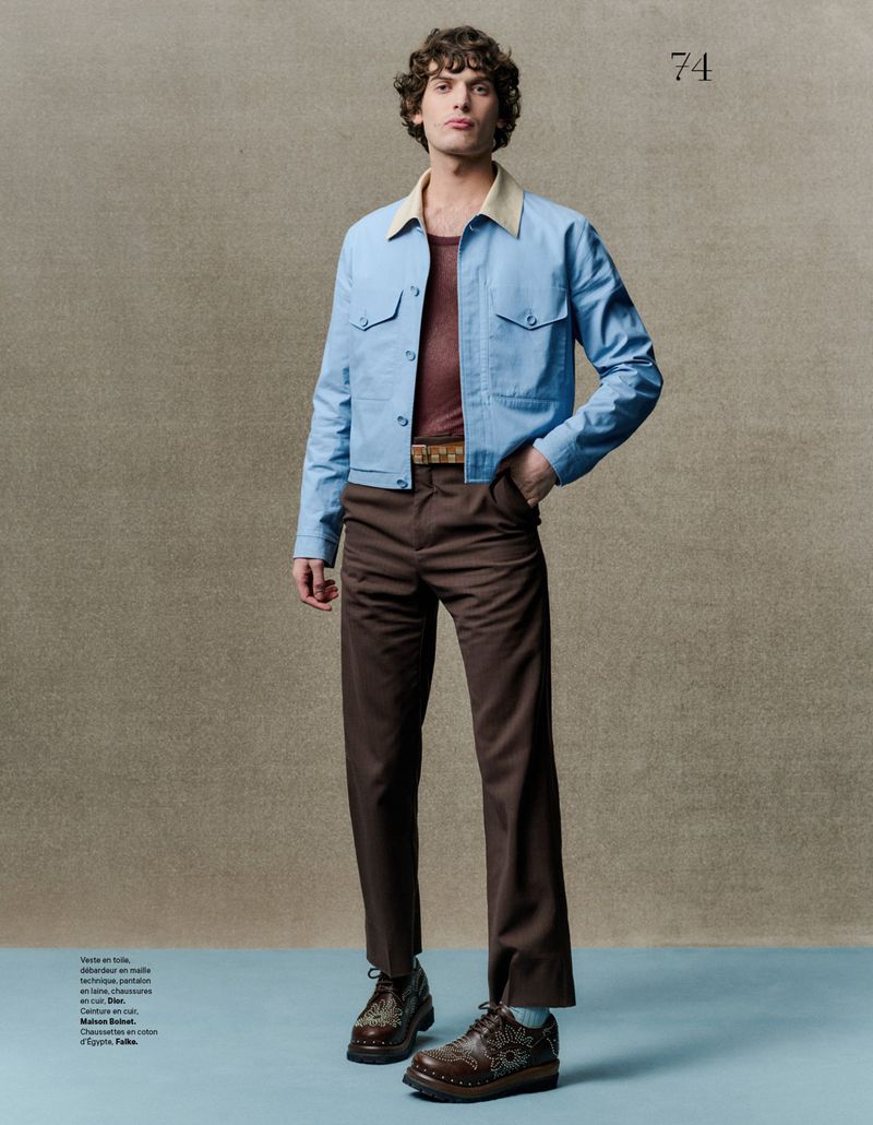 Man poses in blue jacket, brown pants, and patterned shoes in a studio setting. Full shot of a young adult in stylish attire.