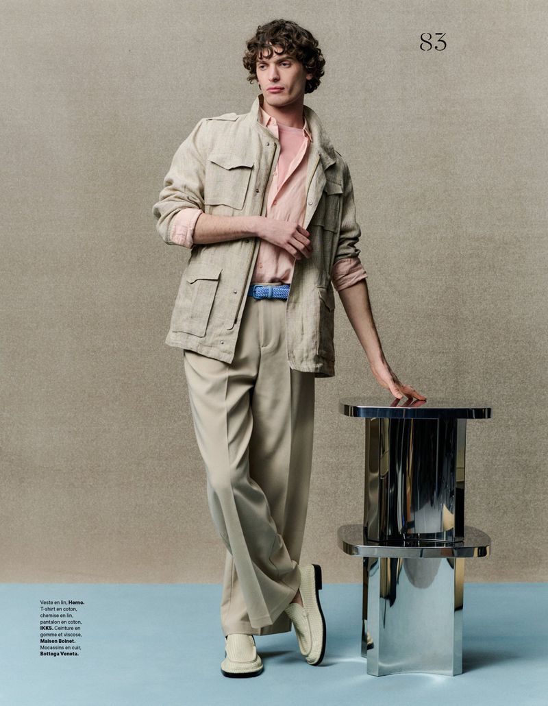 A male model poses in neutral tones wearing a safari jacket, light pink shirt, and beige trousers, leaning on a polished metal table.