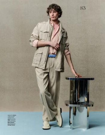 A male model poses in neutral tones wearing a safari jacket, light pink shirt, and beige trousers, leaning on a polished metal table.