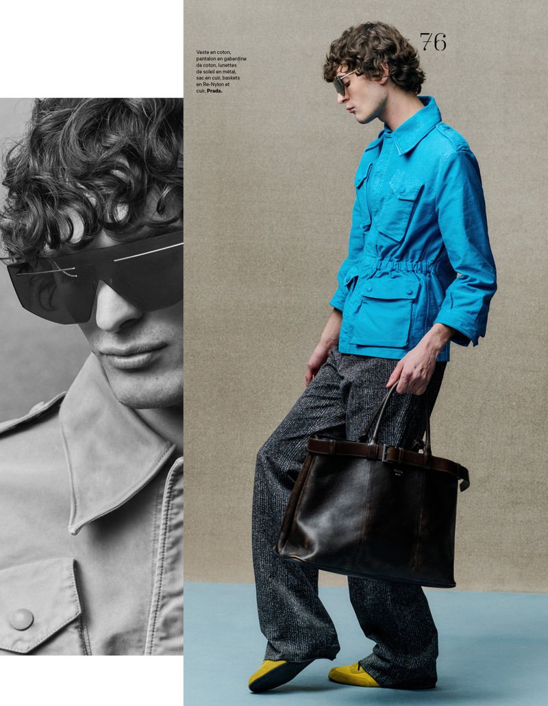 Fashion shot of a young adult male model wearing a blue jacket, gray trousers, sunglasses, and carrying a large brown leather bag.