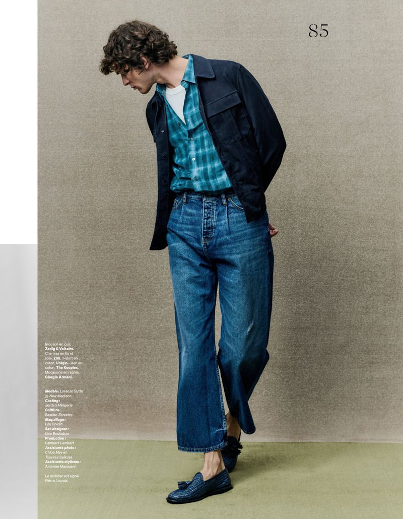A cool toned medium shot captures a slender adult male in a plaid shirt, jeans, and a dark jacket, set against a neutral backdrop.