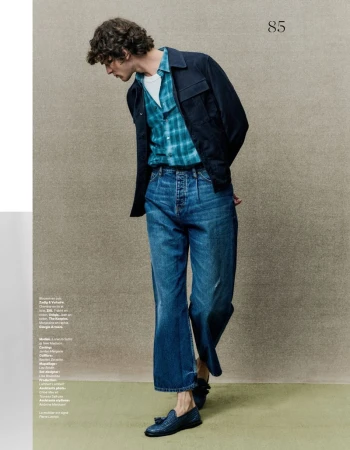 A cool toned medium shot captures a slender adult male in a plaid shirt, jeans, and a dark jacket, set against a neutral backdrop.