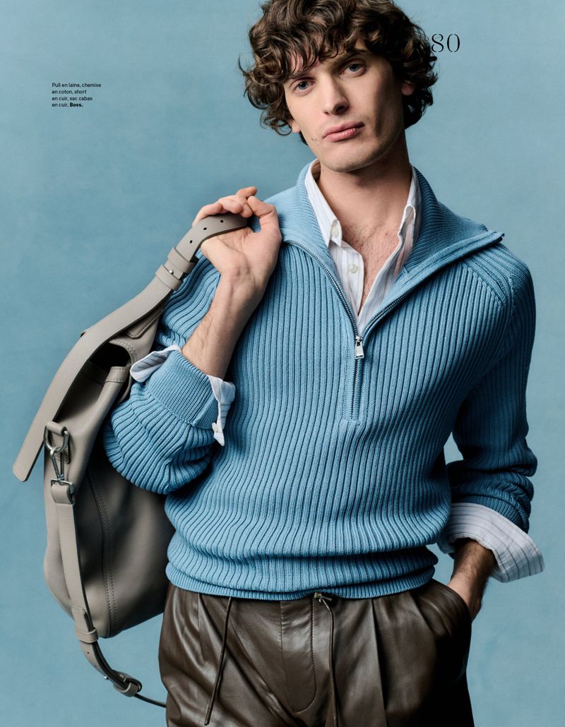 Portrait of a man with curly hair wearing a blue sweater and brown pants, carrying a gray messenger bag on his shoulder.