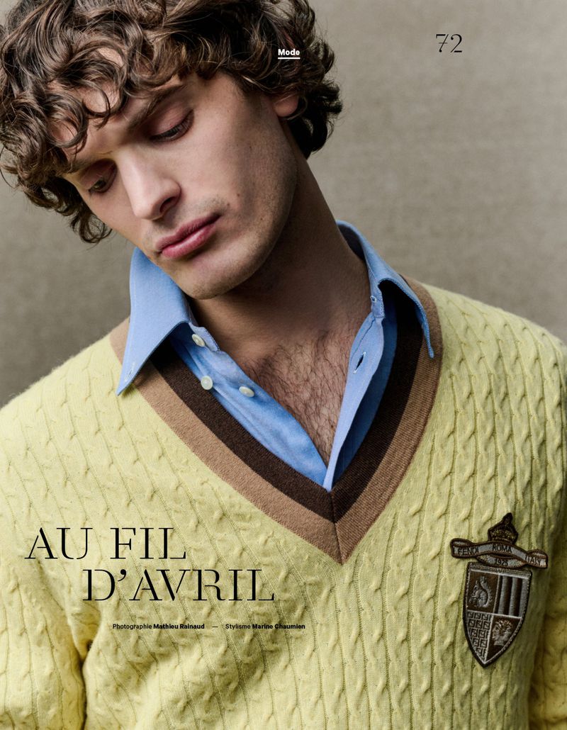 Editorial fashion shot of a young adult man with curly hair wearing a yellow cable-knit sweater over a blue collared shirt indoors.