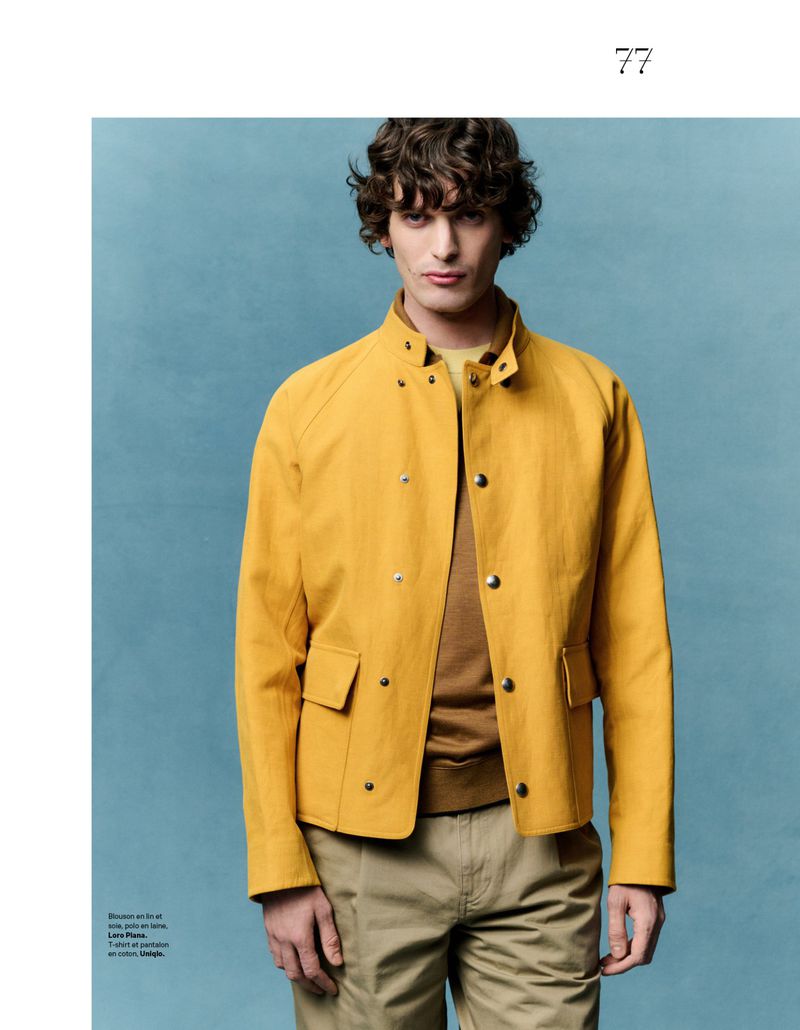 A young adult male model poses in a studio with a blue backdrop, wearing a yellow collared jacket, brown shirt, and khaki-colored trousers.
