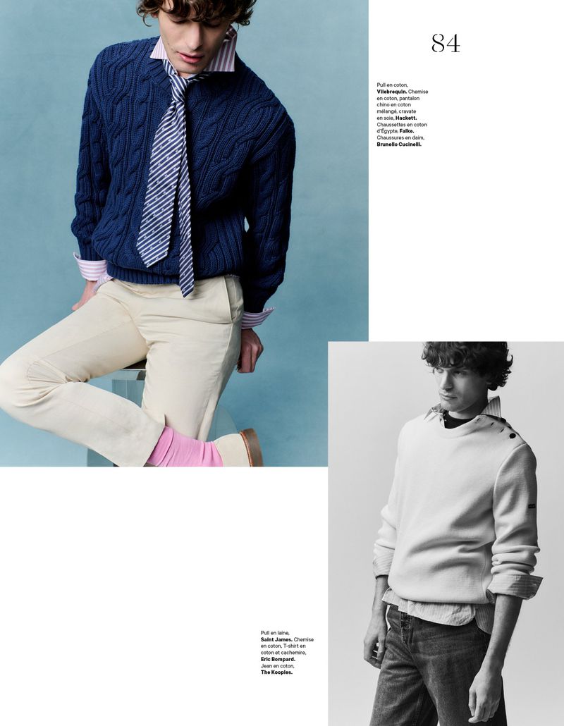 Editorial fashion shot of a young man wearing a navy cable knit sweater, striped shirt and tie, and cream-colored pants, posing indoors.