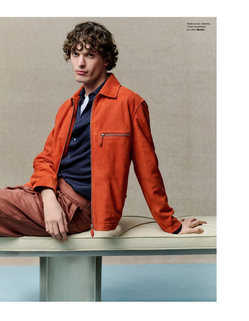 A young adult male poses indoors on a modern bench, wearing a coral jacket, navy button-down shirt, and coral pants in a fashion editorial.