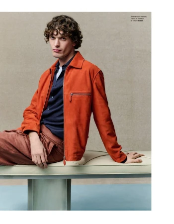 A young adult male poses indoors on a modern bench, wearing a coral jacket, navy button-down shirt, and coral pants in a fashion editorial.