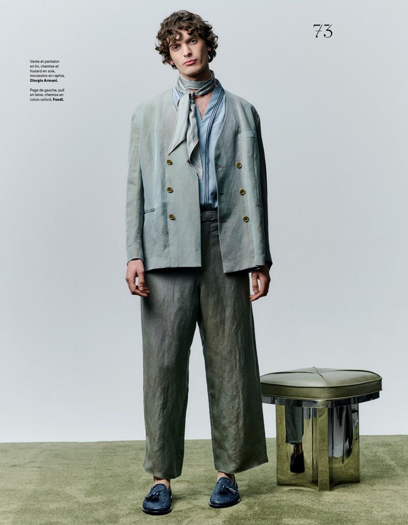 A curly-haired man in a light blue suit and scarf stands next to a green stool in a studio with a light gray backdrop.