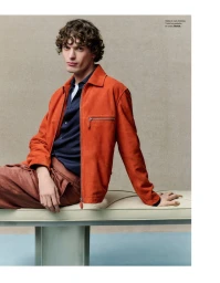 Male model wearing a rust orange leather jacket and dark shirt, seated on a padded bench in a studio setting.