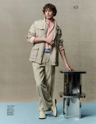 Model leaning on a metallic pedestal wearing a beige utility jacket, pink shirt, and trousers