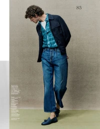 Full-length editorial image of a male model wearing blue denim trousers, a plaid shirt, and a dark jacket against a textured wall.