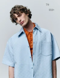 Young man with curly brown hair wearing a light blue button-down shirt and orange polo shirt against a grey background