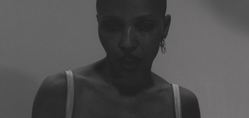 Monochromatic portrait of a woman with a shaved head, wearing an earring, and a white top against a neutral gray background.