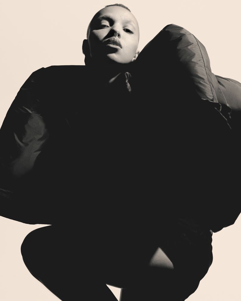 A person with a shaved head poses in a black jacket against a cream-colored backdrop, featuring an eye-level angle and portrait orientation.