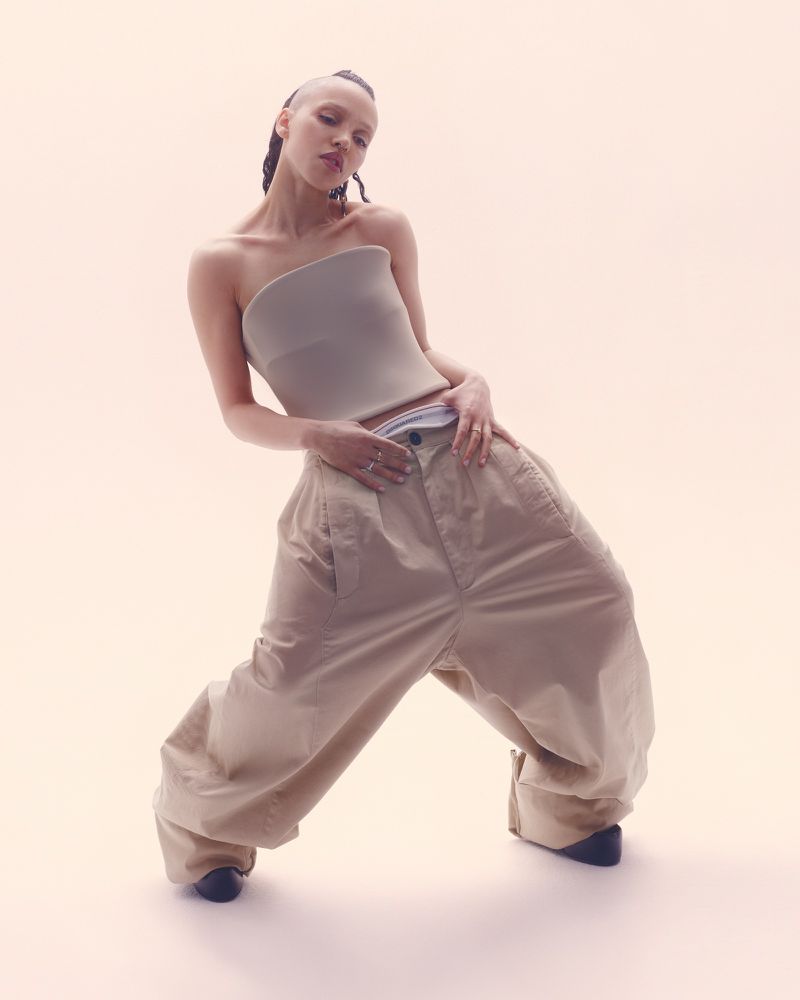 A stylish adult female poses in a light studio wearing a tube top, oversized pants, and dark footwear, photographed from a low angle.