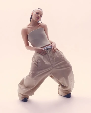 A stylish adult female poses in a light studio wearing a tube top, oversized pants, and dark footwear, photographed from a low angle.