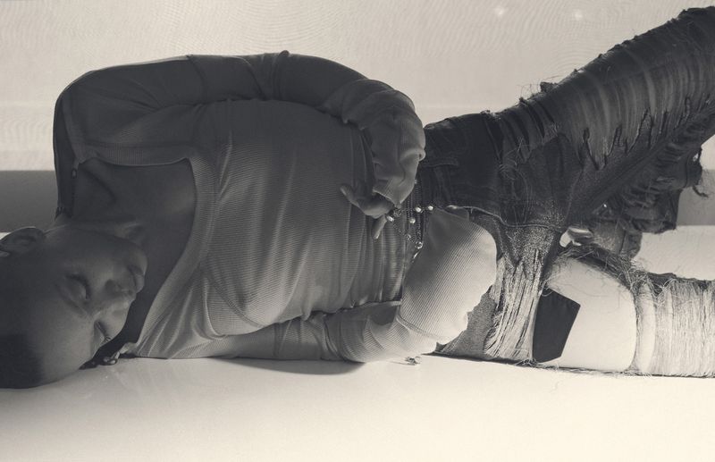Monochrome artistic shot of a person lying on a white surface wearing casual clothing with tattered fabric details, expressing a quiet mood.
