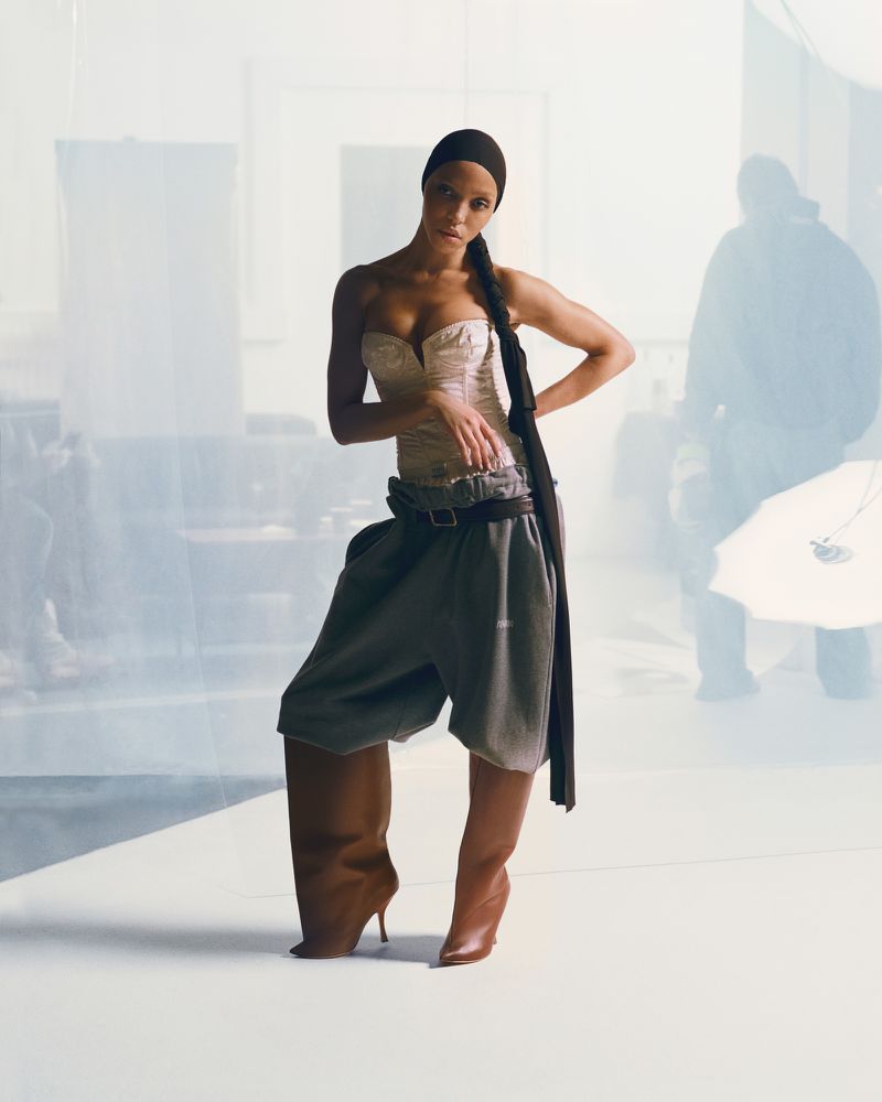 A solo female fashion model poses in a studio wearing a pale bustier top, loose grey bottoms, brown belt, and matching brown heeled boots.