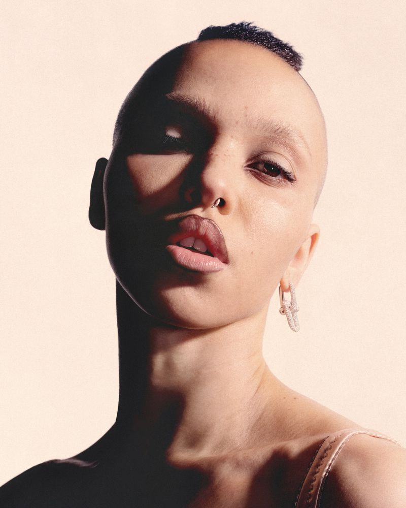 Close-up portrait of a female with a shaved head, light skin, and a nose ring, wearing earrings, against a soft peach background with light shadows.