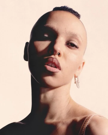 Close-up portrait of a female with a shaved head, light skin, and a nose ring, wearing earrings, against a soft peach background with light shadows.