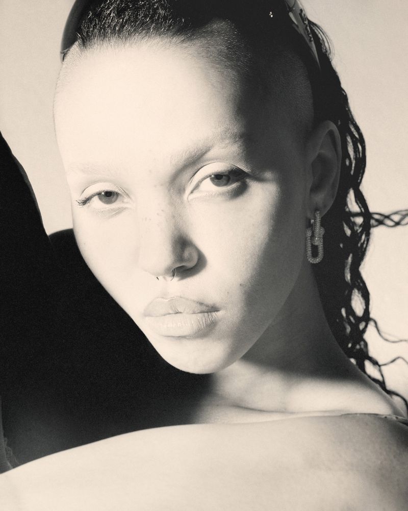 Close-up portrait of a Black woman with shaved sides and cornrow braids, wearing hoop earrings and a black top against a neutral background.