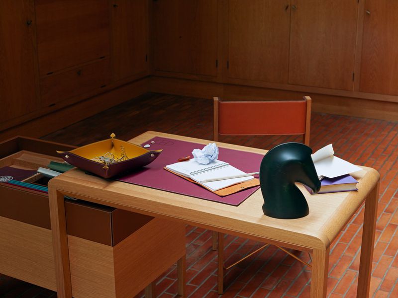 Office interior featuring a wooden desk with a dark green chess piece, notebooks, a leather tray, and an open drawer, in warm lighting.