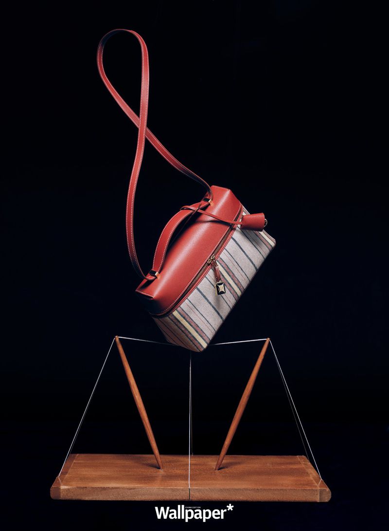 Fashion still life featuring a brown leather and striped fabric handbag suspended above a wooden display stand against a black background.