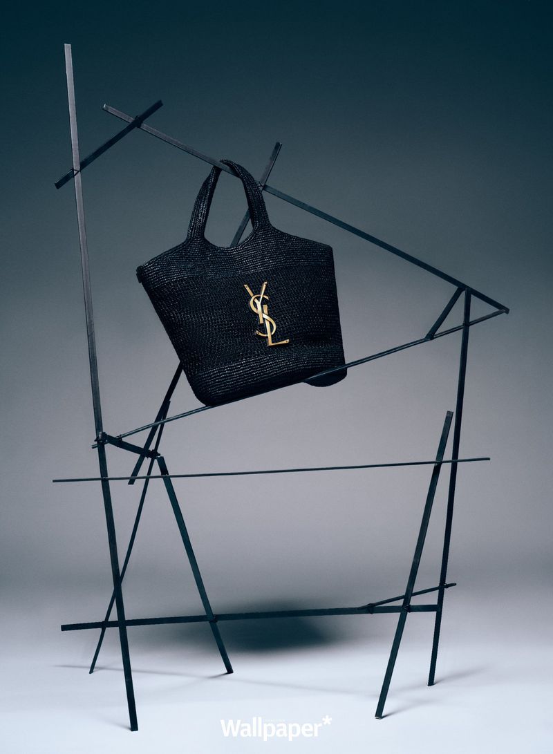 Black woven tote bag by Saint Laurent, displayed on a modernistic black metal sculpture against a dark gray background.