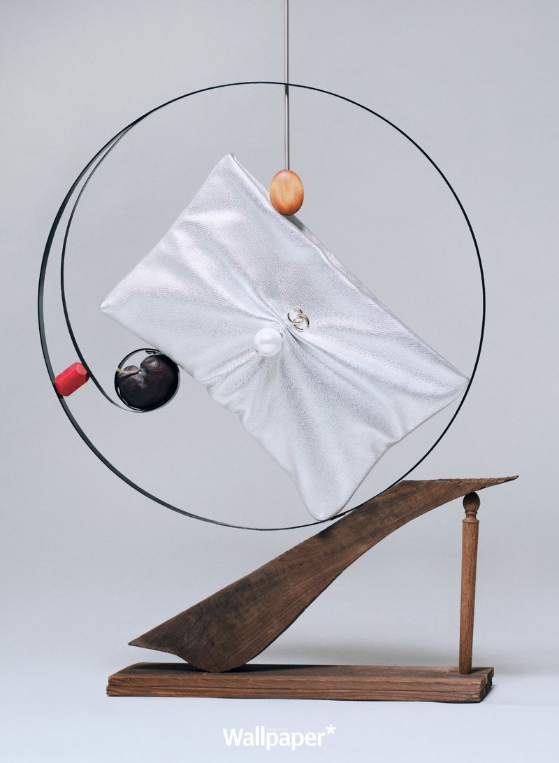 Abstract sculpture featuring a white, fabric-covered object, suspended within a circular frame, balanced on a wooden support against a grey backdrop.
